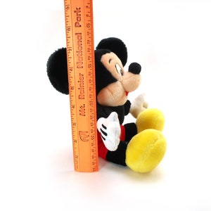 K&ouml;nnte beinhalten: Ein schwarzes und rotes Pl&uuml;sch-Mickey-Mouse-Spielzeug, das auf einer wei&szlig;en Oberfl&auml;che sitzt. Das Spielzeug ist etwa 25 cm gro&szlig; und hat ein gelbes Lineal daneben.