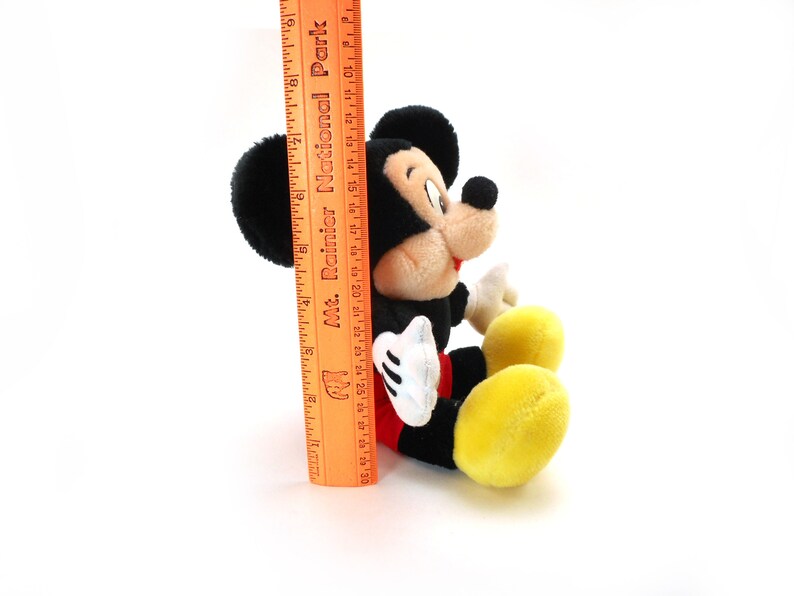 K&ouml;nnte beinhalten: Ein schwarzes und rotes Pl&uuml;sch-Mickey-Mouse-Spielzeug, das auf einer wei&szlig;en Oberfl&auml;che sitzt. Das Spielzeug ist etwa 25 cm gro&szlig; und hat ein gelbes Lineal daneben.