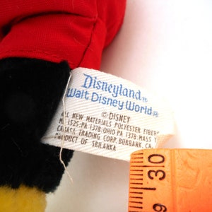 K&ouml;nnte beinhalten: Ein rotes und schwarzes Pl&uuml;sch-Mickey-Mouse-Spielzeug mit einem gelben Gesicht. Das Spielzeug hat ein Etikett mit der Aufschrift "Disneyland Walt Disney World" und enth&auml;lt Informationen &uuml;ber die Materialien und den Hersteller.
