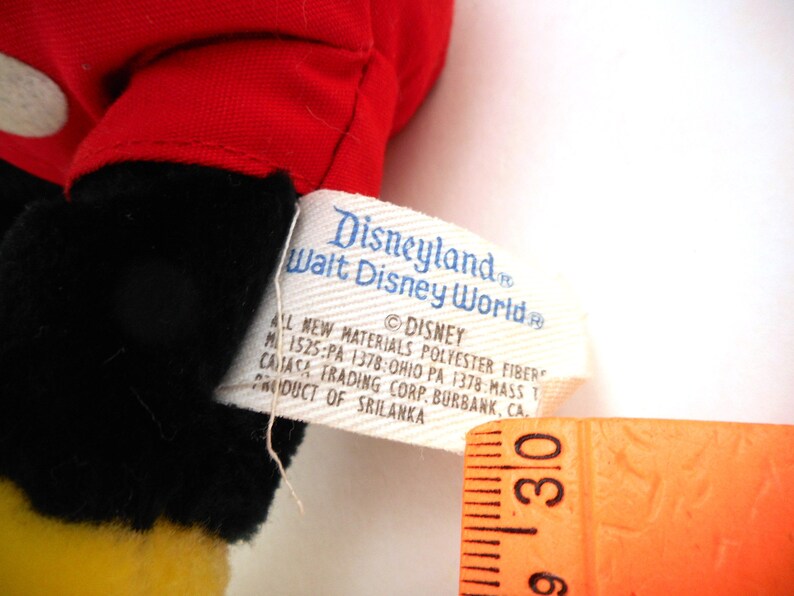 K&ouml;nnte beinhalten: Ein rotes und schwarzes Pl&uuml;sch-Mickey-Mouse-Spielzeug mit einem gelben Gesicht. Das Spielzeug hat ein Etikett mit der Aufschrift "Disneyland Walt Disney World" und enth&auml;lt Informationen &uuml;ber die Materialien und den Hersteller.