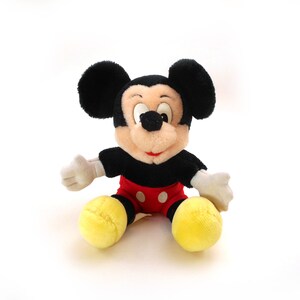 K&ouml;nnte beinhalten: Ein schwarzes und rotes Pl&uuml;sch-Mickey-Mouse-Spielzeug, das auf einem wei&szlig;en Hintergrund sitzt. Das Spielzeug hat gro&szlig;e schwarze Ohren, eine schwarze Nase und gelbe Schuhe.