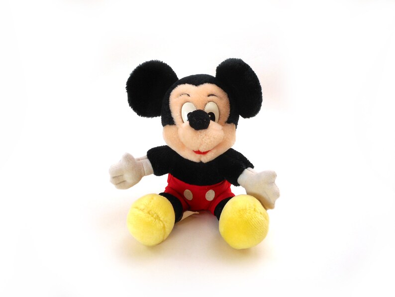 K&ouml;nnte beinhalten: Ein schwarzes und rotes Pl&uuml;sch-Mickey-Mouse-Spielzeug, das auf einem wei&szlig;en Hintergrund sitzt. Das Spielzeug hat gro&szlig;e schwarze Ohren, eine schwarze Nase und gelbe Schuhe.
