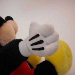 K&ouml;nnte beinhalten: Ein Pl&uuml;sch-Mickey-Mouse-Spielzeug mit schwarzem Fell, einem roten Shirt und einer gelben Hose. Das Spielzeug hat wei&szlig;e Handschuhe mit schwarzen N&auml;hten.