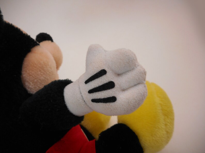 K&ouml;nnte beinhalten: Ein Pl&uuml;sch-Mickey-Mouse-Spielzeug mit schwarzem Fell, einem roten Shirt und einer gelben Hose. Das Spielzeug hat wei&szlig;e Handschuhe mit schwarzen N&auml;hten.