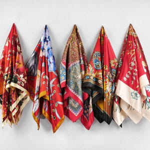 May include: Five silk scarves with vibrant floral and paisley patterns hang against a white wall. The scarves feature a range of colors, including red, blue, gold, and black, with intricate designs and contrasting borders. Each scarf is folded and displayed to showcase its unique pattern.