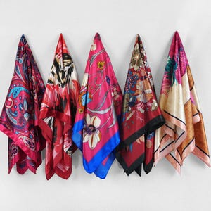 May include: Five silk scarves in shades of pink, red, and cream hang against a white wall. Each scarf features a unique pattern, including paisley, floral, and abstract designs. The scarves are square and appear to be made of a lightweight, flowing fabric.