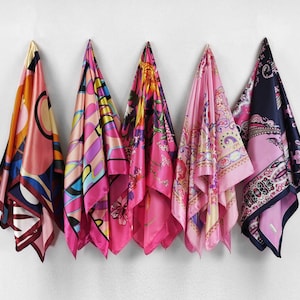 May include: Five colorful silk scarves hanging against a white wall. The scarves feature vibrant patterns in shades of pink, orange, blue, and purple. Each scarf has a unique design, showcasing a variety of abstract and floral motifs.