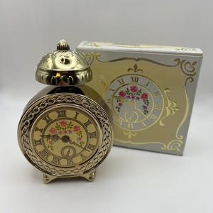 May include: A gold-colored, vintage-style clock with a round face featuring Roman numerals and a floral design. The clock has a matching lid and sits on small feet. A decorative box with a clock face design is behind the clock.
