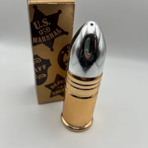 May include: A gold-colored bullet-shaped bottle with a silver top, next to a gold box. The box has the words "U.S. MARSHAL" and "DEPUTY SHERIFF" printed on it. The bottle is likely aftershave or cologne.