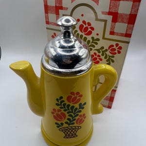 Puede incluir: Una cafetera de cerámica amarilla con tapa plateada y diseño floral. La cafetera tiene un pico y un asa. La caja detrás de la cafetera tiene un patrón de cuadros rojos y blancos y detalles florales.