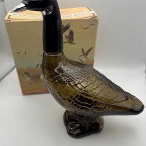 May include: A vintage aftershave bottle in the shape of a goose, featuring a black head and neck, a silver beak, and a textured brown body. The bottle is on a base, with a box in the background showing a wildlife illustration.
