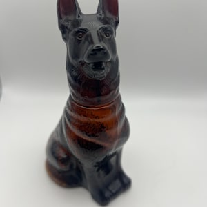 May include: A dark brown glass ornament of a German Shepherd dog in a seated pose. The dog has upright ears and a detailed face. The glass has a glossy finish, with a darker shade on the ears and nose.
