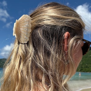May include: A cream-colored hair claw clip shaped like a scallop shell with a pearlescent finish. The clip features a textured, ribbed surface mimicking the natural ridges of a seashell and is used to secure a section of blonde hair in place.