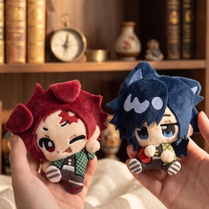 May include: Two plush toys featuring stylized characters with large eyes. One plush has dark red hair and a green and black checkered outfit. The other plush has dark blue hair, cat-like ears, and a green, yellow, and red patterned outfit.