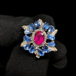 May include: A silver ring featuring a central oval-cut vibrant pink gemstone surrounded by a halo of small clear stones. A secondary outer layer consists of twelve oval-cut blue gemstones arranged in a starburst pattern with clear accents.