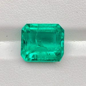 May include: A vivid, emerald-cut gemstone in a deep green shade. The gem has a rectangular shape with angled edges, displaying its facets and internal clarity. The stone is set against a white backdrop, accentuating its colour and brilliance.
