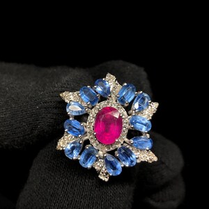 May include: A gemstone ring featuring a large oval ruby center stone surrounded by blue sapphires and small diamonds. The ring has a silver-colored setting and is displayed against a black background.