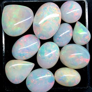 Può includere: Una collezione di opali di varie forme, tra cui ovali e a goccia. Gli opali mostrano un gioco di colori con lampi iridescenti di rosa, verde e blu su una base bianco latte. Le pietre preziose sono disposte su uno sfondo scuro.