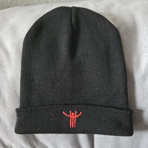 May include: Black knit beanie with a folded brim. The beanie features a small, embroidered orange design on the front. The hat is made of a soft, knitted material.