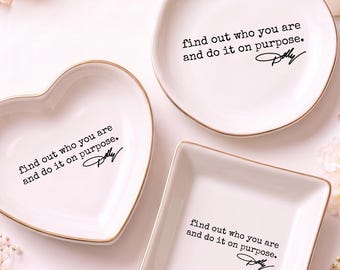 May include: Three white ceramic trinket dishes in heart, round, and square shapes with gold-toned rims. Each dish features the black printed text: find out who you are and do it on purpose. Dolly, in a typewriter-style font centered on the base.
