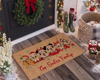 May include: A rectangular tan coir doormat featuring a colorful illustration of Disney characters including Pluto, Donald Duck, Minnie Mouse, Mickey Mouse, Daisy Duck, and Goofy wearing Santa hats, with the red text The Smith Family.
