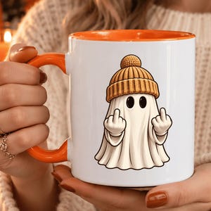 May include: A white ceramic mug with an orange interior and handle, featuring a graphic of a white ghost wearing a tan beanie hat and holding up both middle fingers. The design is centered on the front of the mug, which has a smooth, glossy finish.