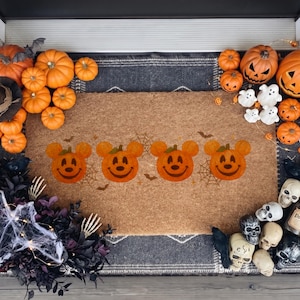 May include: A rectangular tan coir doormat featuring four orange pumpkin icons with mouse-ear shapes and smiling faces. The mat is surrounded by various Halloween decorations, including several orange pumpkins, white ghost figurines, and human skull replicas.