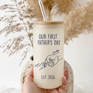 Peut inclure: Un gobelet en verre givré en forme de canette avec un couvercle en bois et une paille transparente. Le texte "OUR FIRST FATHER'S DAY" est imprimé sur le devant, ainsi qu'un dessin d'une main tenant la main d'un bébé, le nom "Liam", le nom "Daddy" et l'année "EST. 2026".