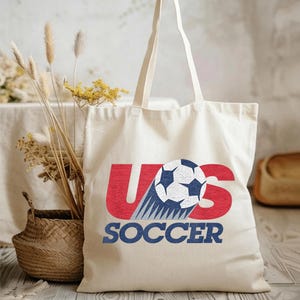 Pode incluir: Uma sacola de lona na cor creme com um design gr&aacute;fico apresentando o texto vermelho US, uma bola de futebol preta e branca substituindo a letra S e o texto azul SOCCER abaixo. A bolsa possui duas al&ccedil;as longas e uma base plana.