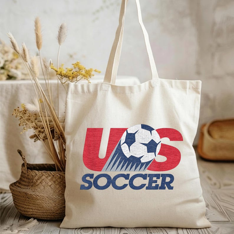Pode incluir: Uma sacola de lona na cor creme com um design gr&aacute;fico apresentando o texto vermelho US, uma bola de futebol preta e branca substituindo a letra S e o texto azul SOCCER abaixo. A bolsa possui duas al&ccedil;as longas e uma base plana.