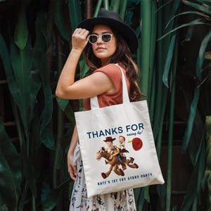 May include: A white canvas tote bag featuring a graphic of Woody riding Bullseye from Toy Story. The text reads THANKS FOR racing by! KATE'S 1ST TOY STORY PARTY in blue lettering. The bag has two shoulder straps and a rectangular shape.