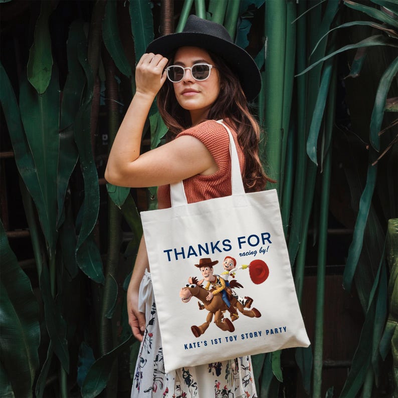 May include: A white canvas tote bag featuring a graphic of Woody riding Bullseye from Toy Story. The text reads THANKS FOR racing by! KATE'S 1ST TOY STORY PARTY in blue lettering. The bag has two shoulder straps and a rectangular shape.