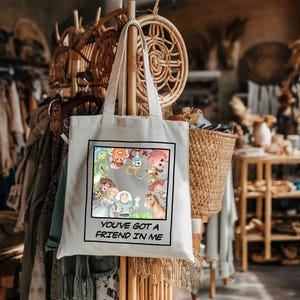 May include: A white canvas tote bag featuring a square graphic print of various animated characters from the Toy Story franchise. Below the colorful character illustration, the text reads, "YOU'VE GOT A FRIEND IN ME" in black capital letters.