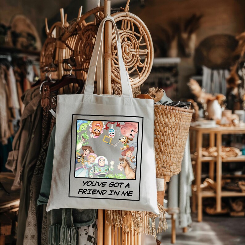 May include: A white canvas tote bag featuring a square graphic print of various animated characters from the Toy Story franchise. Below the colorful character illustration, the text reads, "YOU'VE GOT A FRIEND IN ME" in black capital letters.