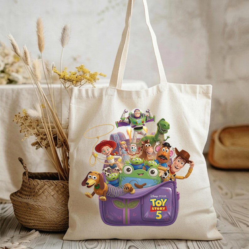 May include: A cream-colored canvas tote bag featuring a colorful graphic of Toy Story characters including Woody, Buzz Lightyear, Jessie, Rex, Slinky Dog, and Mr. Potato Head emerging from a purple backpack with the Toy Story 5 logo printed on it.