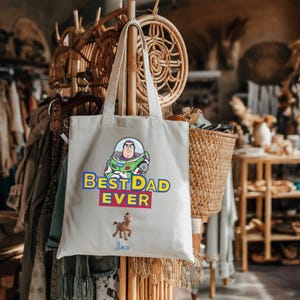 May include: A cream-colored canvas tote bag featuring a graphic of Buzz Lightyear above the blue and yellow text BEST DAD EVER. Below the text is a small illustration of Bullseye the horse and the name Jack written in blue script.