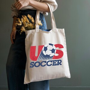 Pode incluir: Uma sacola de lona na cor creme com um design gr&aacute;fico apresentando as letras vermelhas U e S, uma bola de futebol preta e branca substituindo a letra O, e a palavra SOCCER impressa em letras azuis de bloco abaixo do gr&aacute;fico.