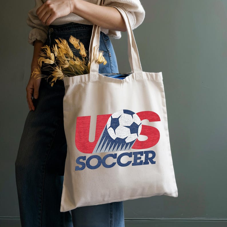 Pode incluir: Uma sacola de lona na cor creme com um design gr&aacute;fico apresentando as letras vermelhas U e S, uma bola de futebol preta e branca substituindo a letra O, e a palavra SOCCER impressa em letras azuis de bloco abaixo do gr&aacute;fico.