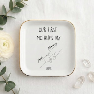 May include: A white square ceramic tray with gold edging. The tray has the text "OUR FIRST MOTHER'S DAY" in black, a line drawing of hands, and the names "Jack" and "Mommy". The year "2026" is also printed on the tray.