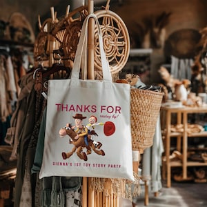 May include: A white canvas tote bag featuring a graphic of Woody and Jessie from Toy Story riding Bullseye. The text reads "THANKS FOR racing by!" and "SIENNA'S 1ST TOY STORY PARTY" in dark pink lettering. The bag has two long shoulder straps.