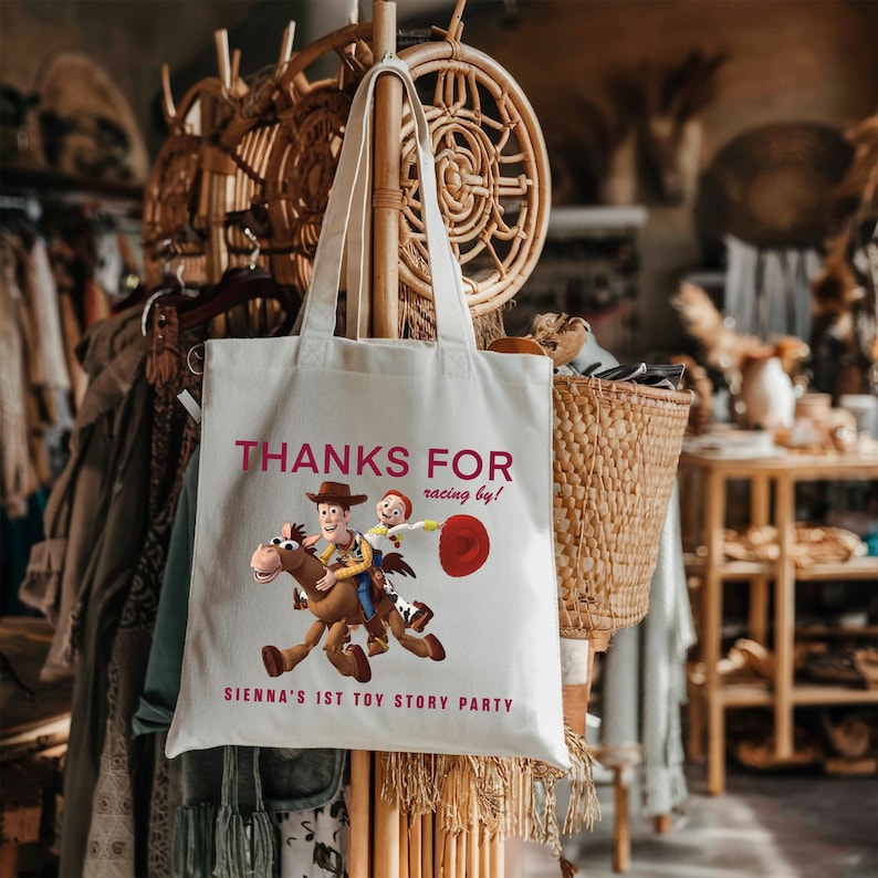 May include: A white canvas tote bag featuring a graphic of Woody and Jessie from Toy Story riding Bullseye. The text reads "THANKS FOR racing by!" and "SIENNA'S 1ST TOY STORY PARTY" in dark pink lettering. The bag has two long shoulder straps.