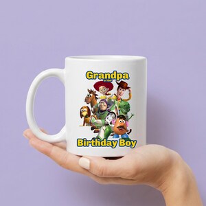 May include: A white ceramic mug featuring a colorful graphic of Toy Story characters including Woody, Buzz Lightyear, Jessie, and Mr. Potato Head. The text "Grandpa of the Birthday Boy" is printed in blue and yellow letters above and below the characters.