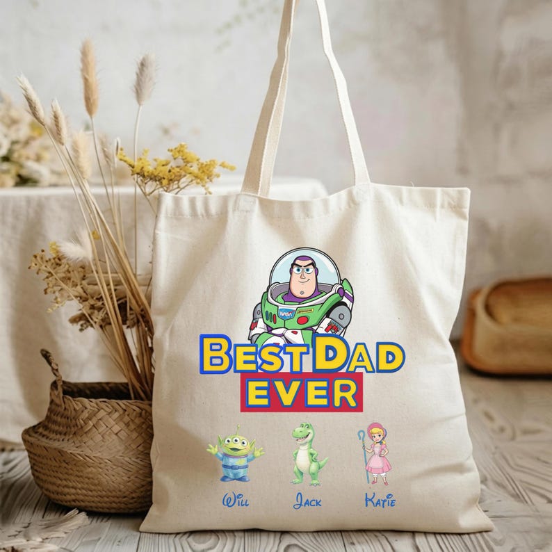 May include: A cream-colored canvas tote bag featuring a graphic of Buzz Lightyear above the text BEST DAD EVER. Below the text are three small character illustrations of an alien, a dinosaur, and a shepherdess with the names Will, Jack, and Katie.