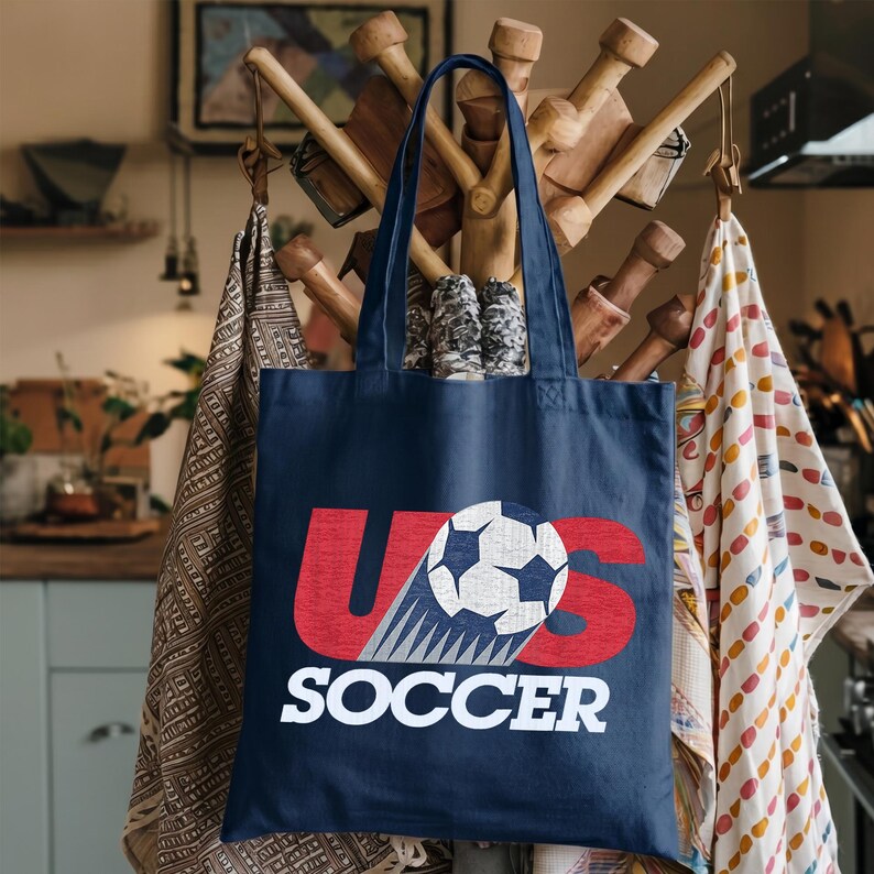 Pode incluir: Uma sacola azul-marinho com um design gr&aacute;fico apresentando as letras vermelhas U e S, uma bola de futebol branca e preta substituindo a letra O, e a palavra SOCCER impressa em letras mai&uacute;sculas brancas abaixo do gr&aacute;fico na frente.