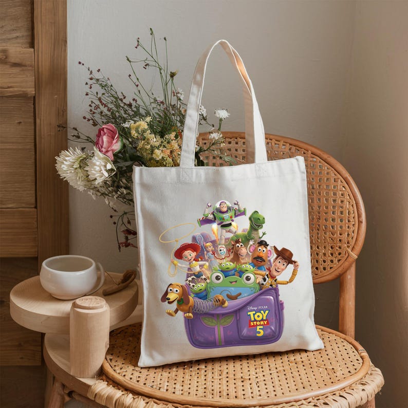May include: A white canvas tote bag featuring a colorful graphic of Toy Story characters including Woody, Buzz Lightyear, Jessie, and Slinky Dog emerging from a purple backpack with the text Disney Pixar Toy Story 5 printed on the front.
