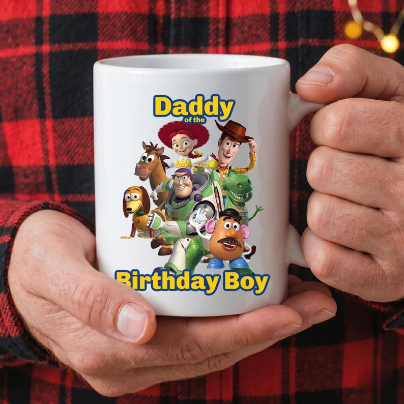 May include: A white ceramic mug featuring a colorful graphic of Toy Story characters including Woody, Buzz Lightyear, Jessie, Bullseye, Rex, Slinky Dog, and Mr. Potato Head, with the blue text Daddy of the Birthday Boy printed on the front.