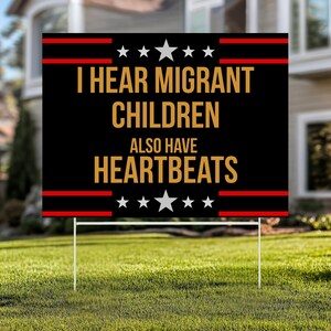 May include: A black yard sign with the text "I HEAR MIGRANT CHILDREN ALSO HAVE HEARTBEATS" in gold lettering. The sign has red stripes and white stars at the top and bottom, and is displayed on a green lawn.