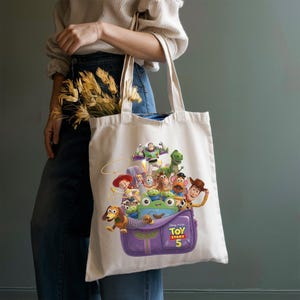 May include: A cream-colored canvas tote bag featuring a colorful graphic print of Toy Story characters including Woody, Buzz Lightyear, Jessie, Rex, and Mr. Potato Head emerging from a purple backpack with the text Toy Story 5 printed on the front.