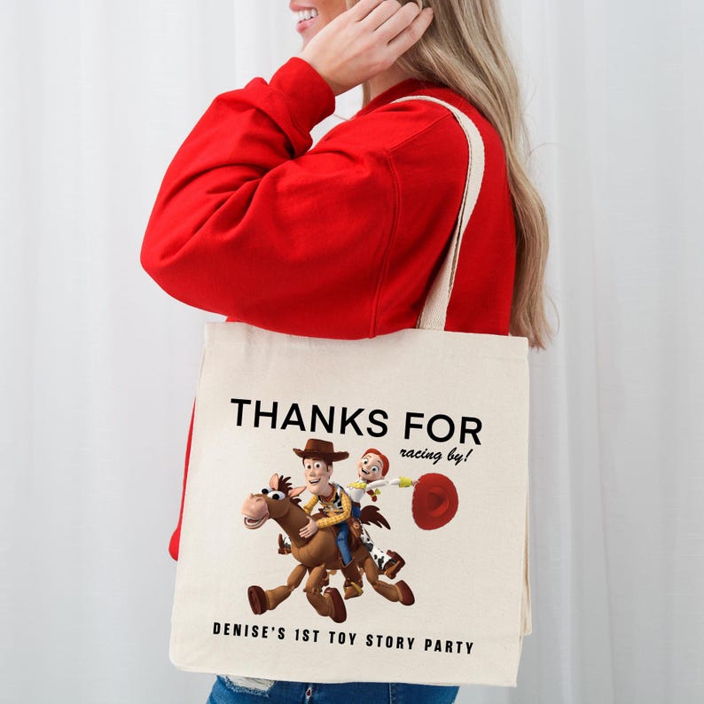 May include: A cream-coloured canvas tote bag featuring a graphic of Woody riding Bullseye from Toy Story. The text reads THANKS FOR racing by! and DENISE'S 1ST TOY STORY PARTY in black lettering. The bag has two shoulder straps and a rectangular shape.