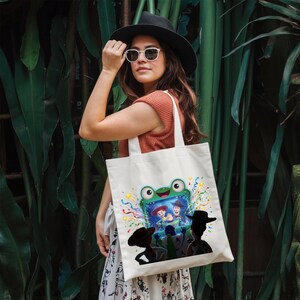 May include: A white canvas tote bag featuring a colorful graphic of a green frog-shaped television screen displaying animated characters, surrounded by confetti. The bag has two sturdy shoulder straps and is made of a durable fabric material.